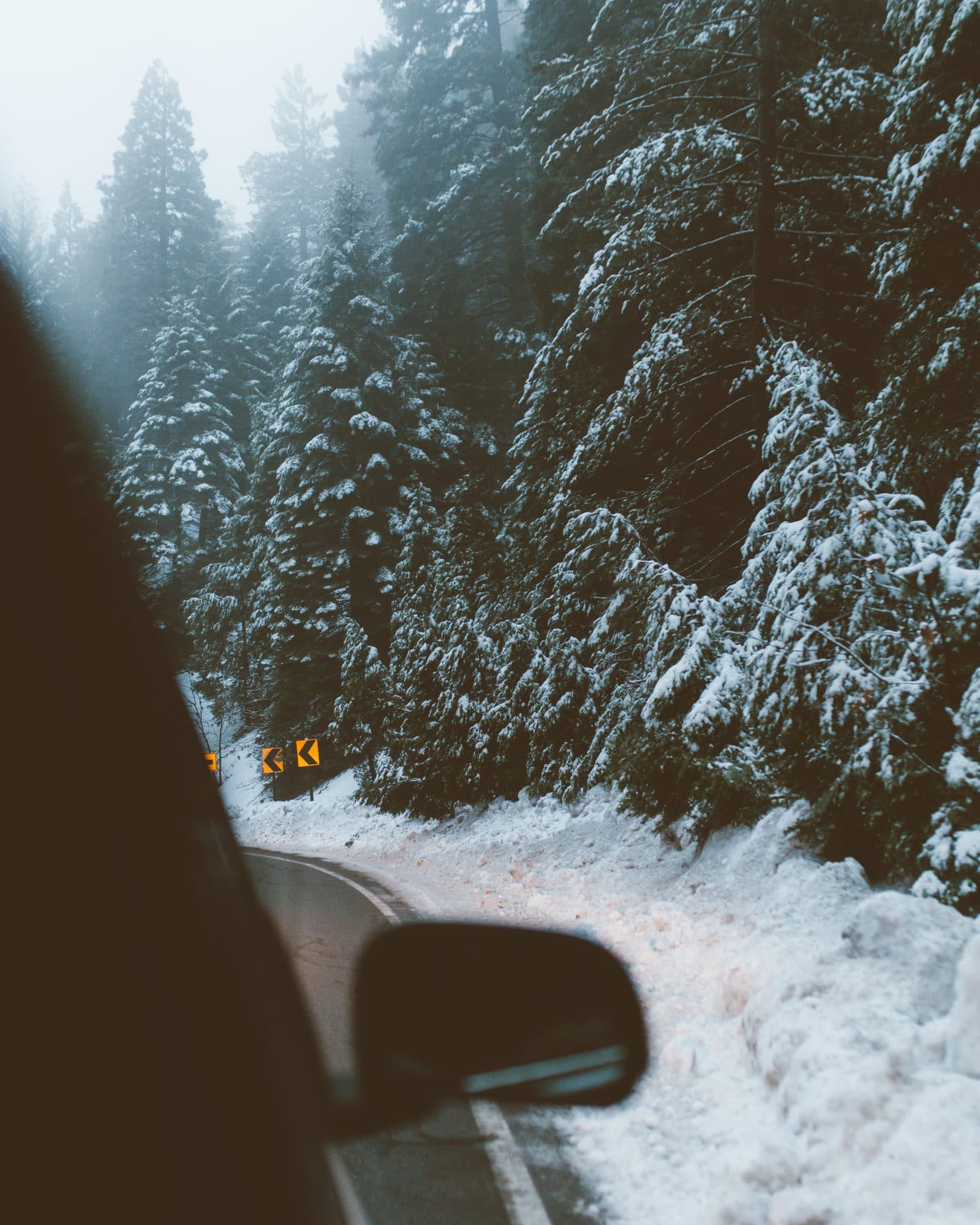 A snow-lined mountain road through tall pines, seen past a car mirror near Lake Arrowhead